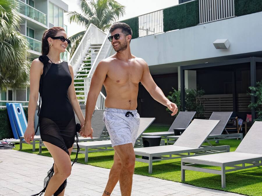 A couple walking on a tiled pathway with lounge chairs and a building in the background.