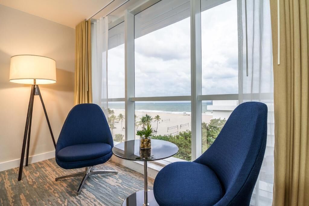 A hotel room with two blue armchairs, a small table with a plant, a floor lamp, and a large window offering a beach view.