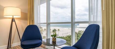 A hotel room with two blue armchairs, a small table with a plant, a floor lamp, and a large window offering a beach view.