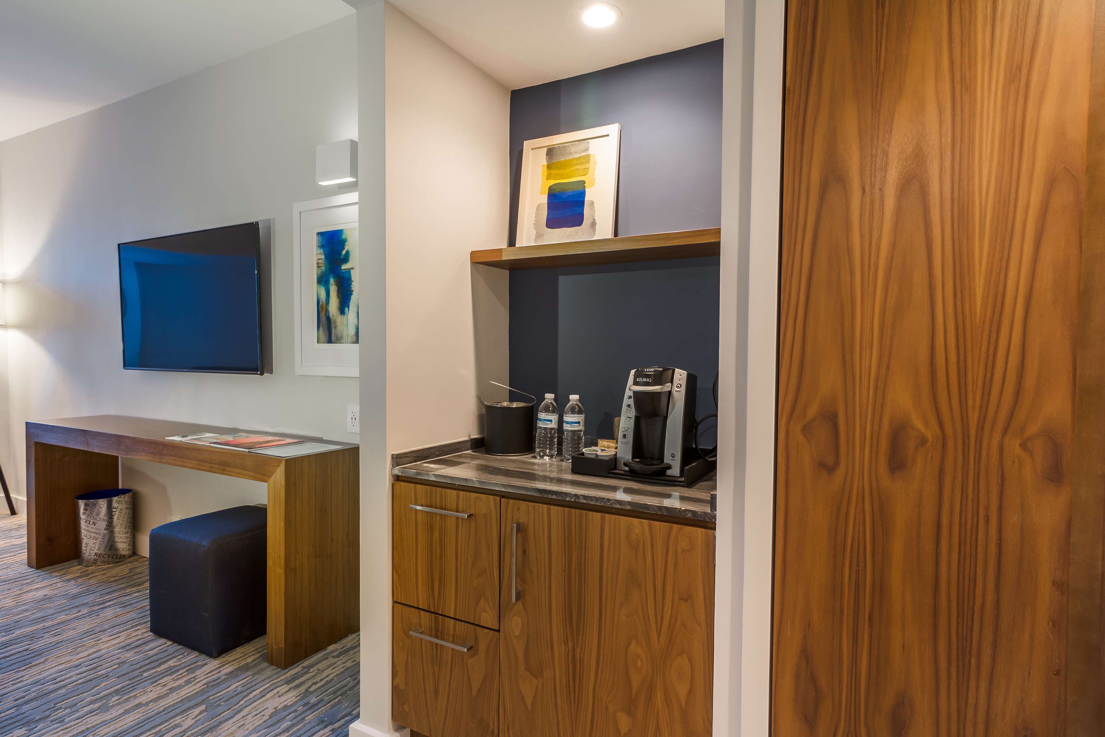 Una kitchenette compacta con cafetera, botellas de agua y una obra de arte enmarcada sobre la encimera.
