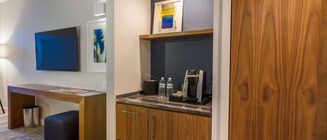 A compact kitchenette with a coffee maker, water bottles, and a framed artwork above the counter.