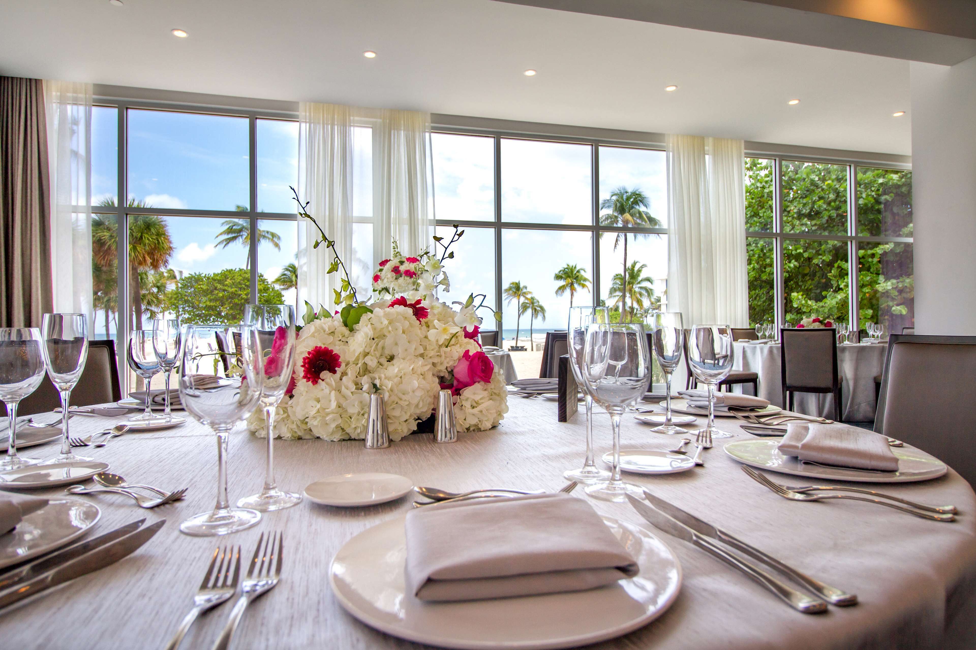 A dining table set for a meal with white plates, silverware, and wine glasses, surrounded by large windows with a view of palm trees.