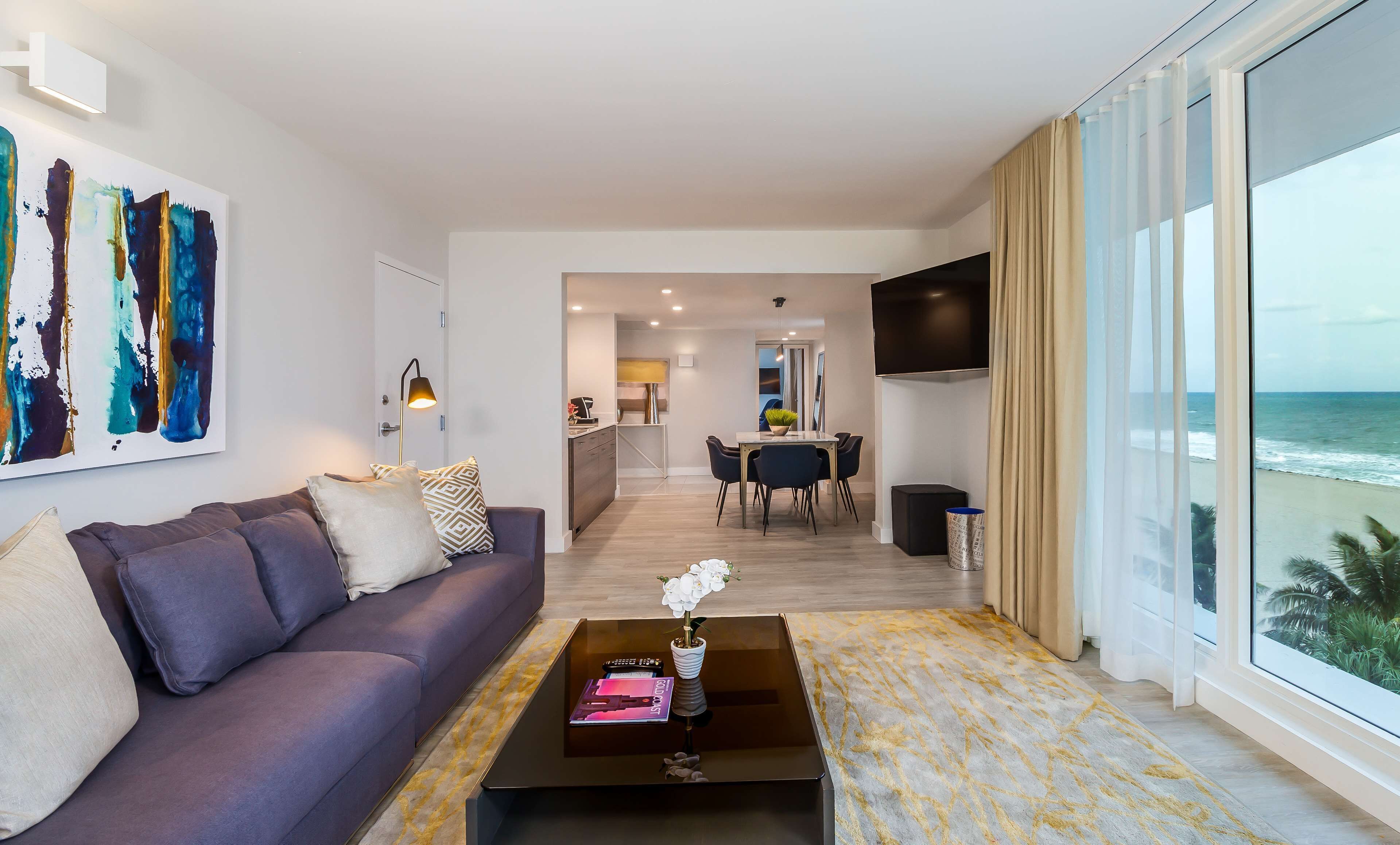 A modern living room with a sofa, coffee table, and artwork. There is a dining area with chairs and a table, and a kitchen in the background. A TV is mounted on the wall, and the room has large windows with a view of the ocean.