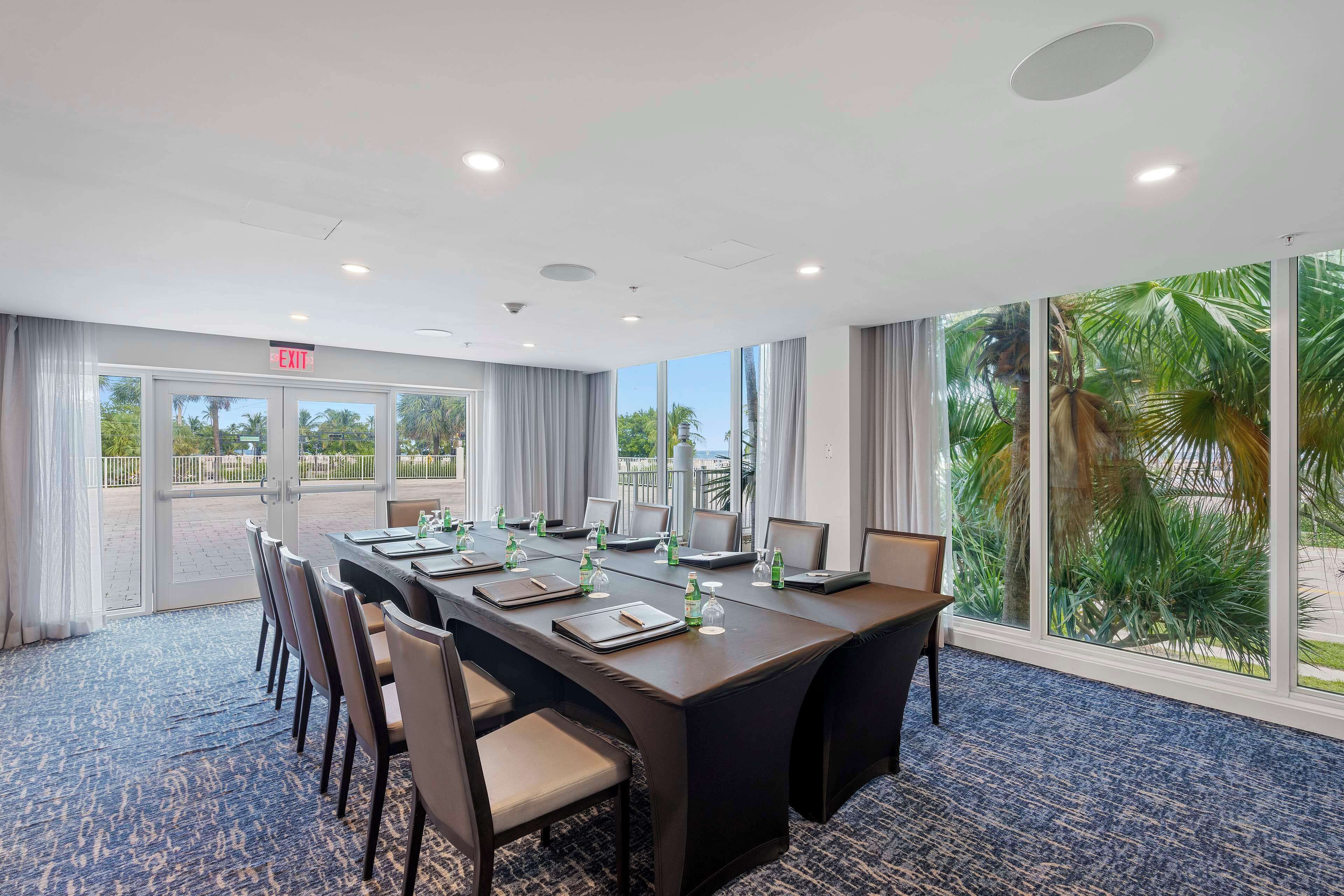 A conference room with a long table, chairs, and a view of palm trees through large windows.