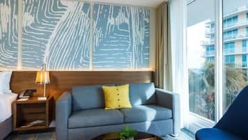 A hotel room with a grey sofa, a blue armchair, a round coffee table with a plant, a wooden nightstand, and a large window offering a view of buildings and palm trees.