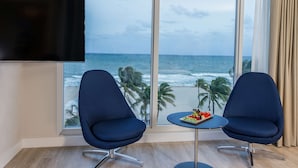 A room with a large window overlooking the ocean, two blue chairs, a small table with fruit, and a mounted TV. - B Ocean Resort Fort Lauderdale Beach (Fort Lauderdale)