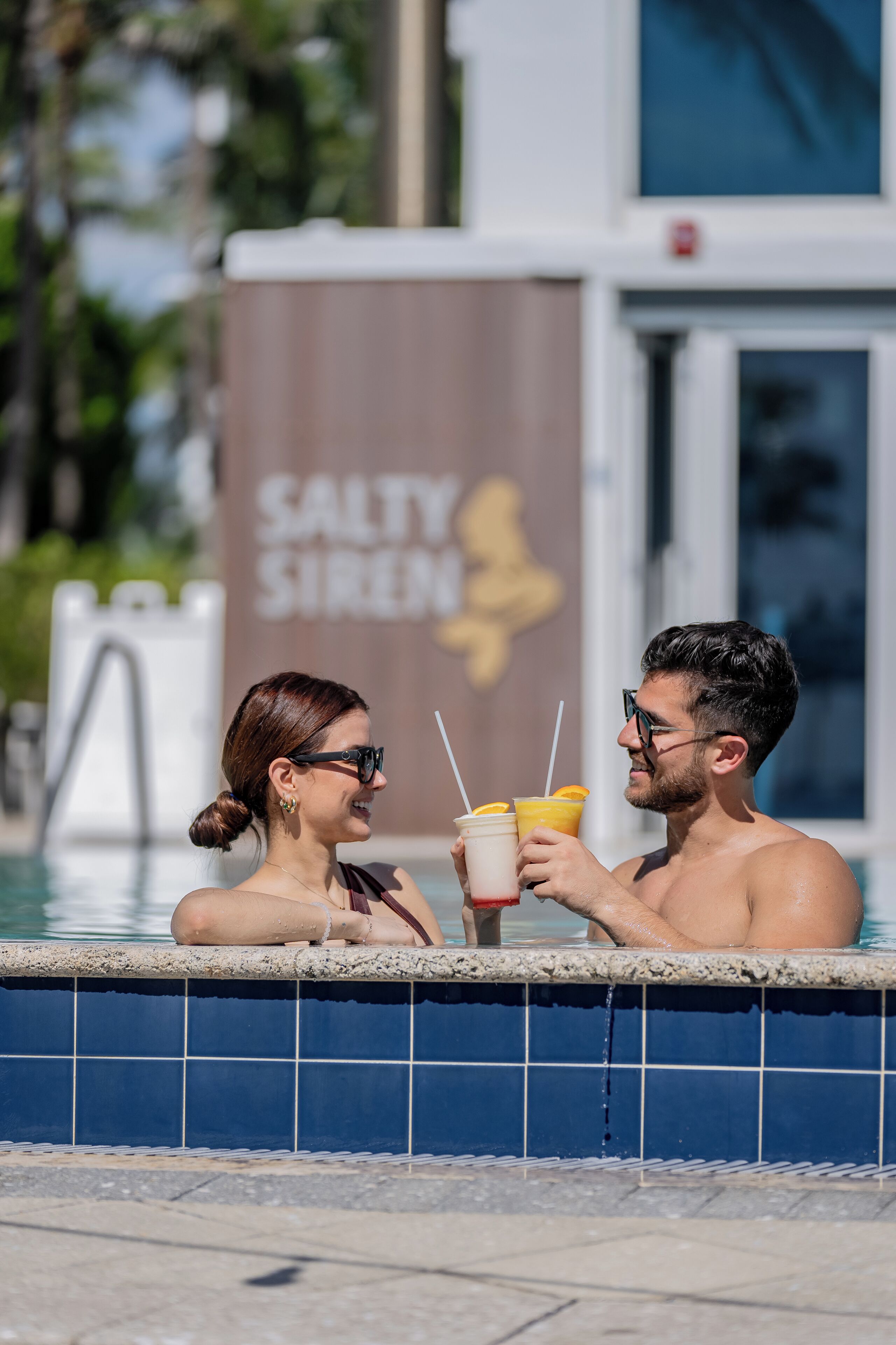 Una pareja en una piscina con bebidas, un edificio con un letrero que dice SALTY SIREN al fondo.