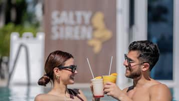 Una pareja en una piscina con bebidas, un edificio con un letrero que dice SALTY SIREN al fondo.