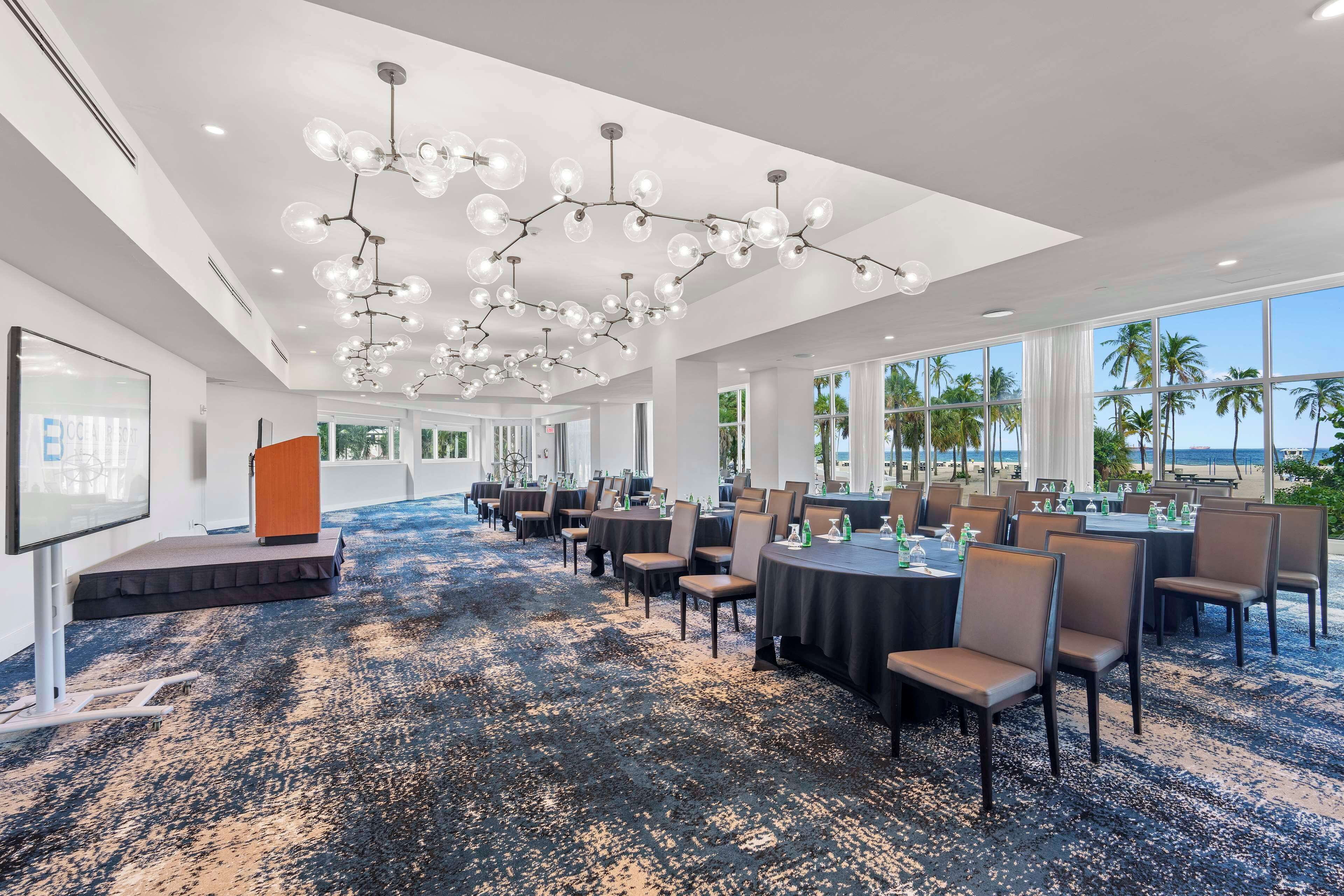 A spacious conference room with a modern chandelier, large windows with a view of palm trees and the ocean, and a carpeted floor.