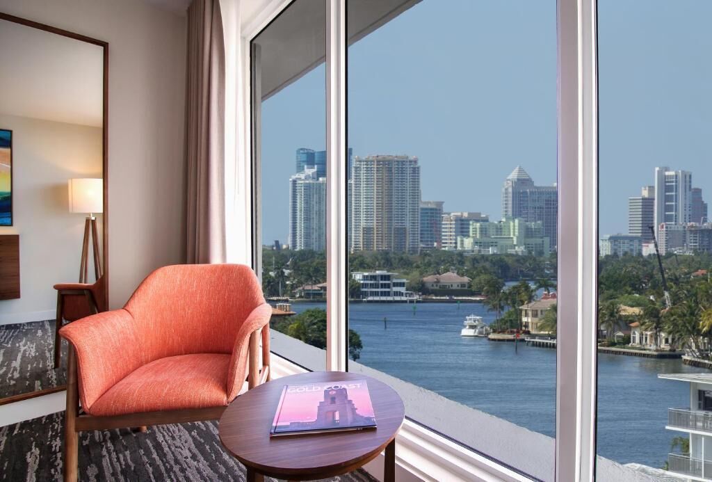 A hotel room with a large window offering a city view, a comfortable orange chair, a small round table, and a magazine titled "SOLD COAST".