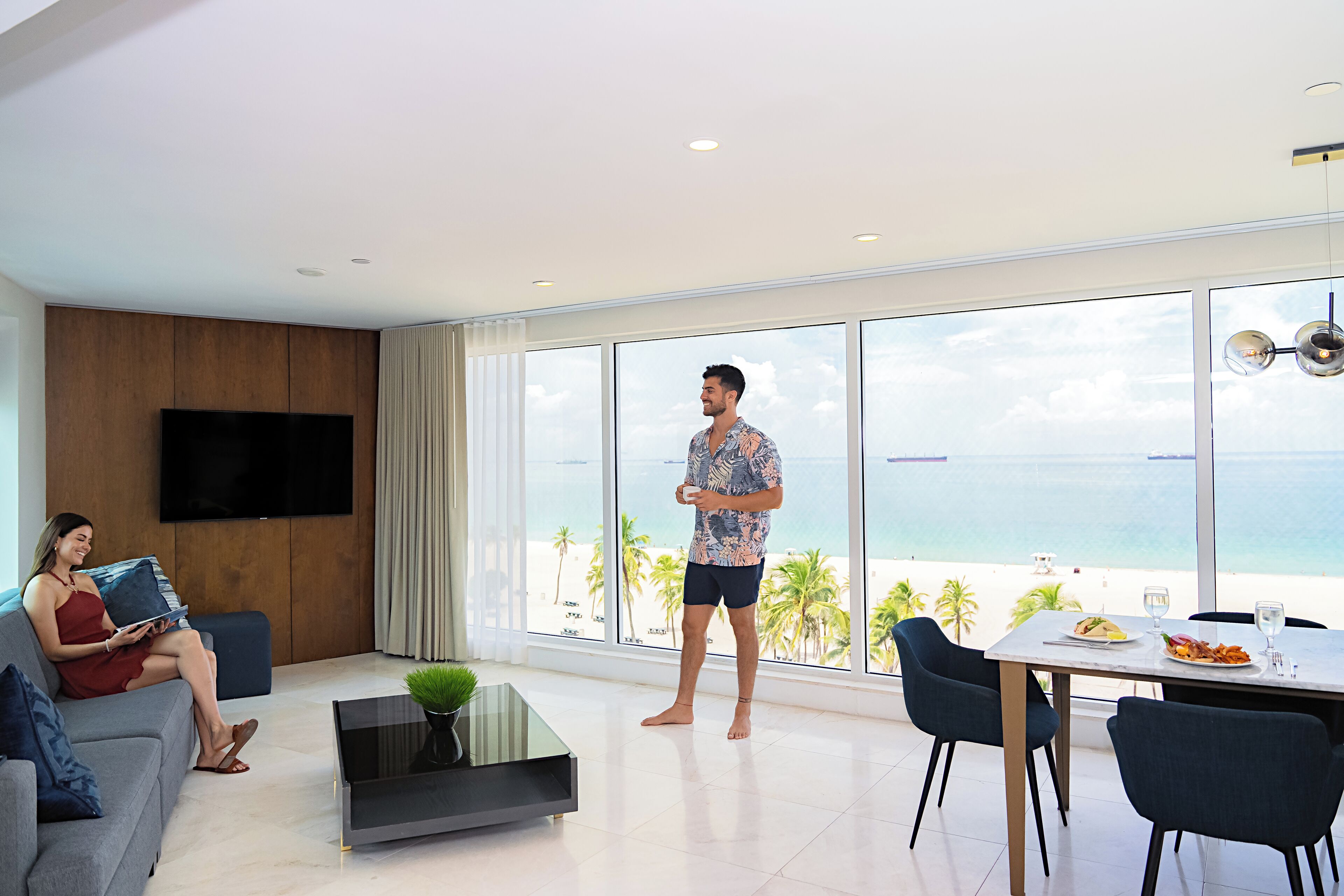 A modern living room with a large window overlooking a beach, a flat-screen TV mounted on the wall, and a dining area with a table and chairs.