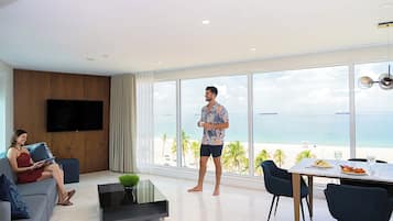 A modern living room with a large window overlooking a beach, a flat-screen TV mounted on the wall, and a dining area with a table and chairs.