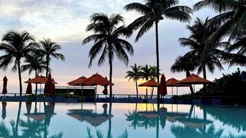 A clear pool with reflections of palm trees and umbrellas, surrounded by lounge chairs and a sunset sky.