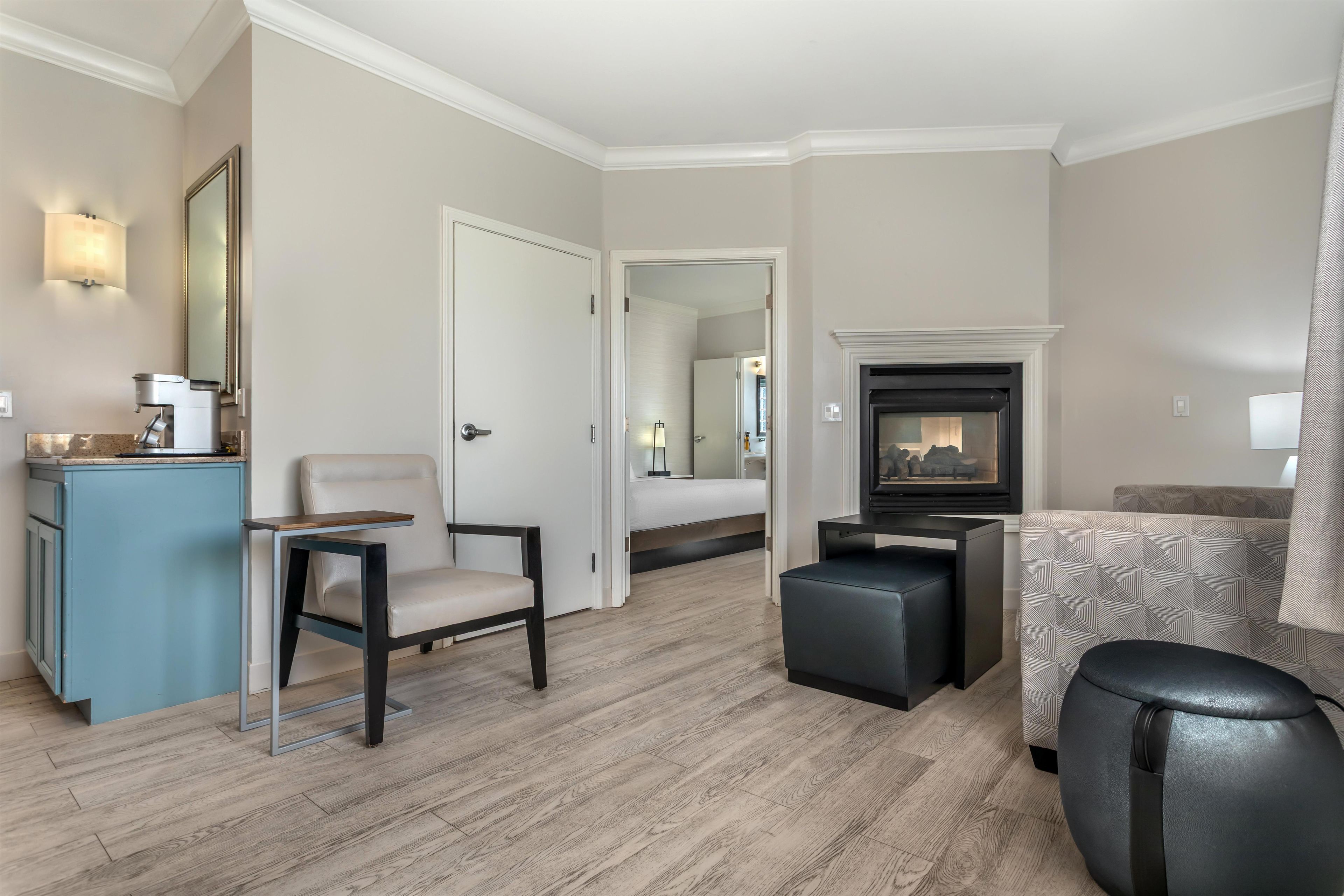 A hotel room with a bed, a seating area with a chair and ottoman, a small table, a fireplace, and a mirror.