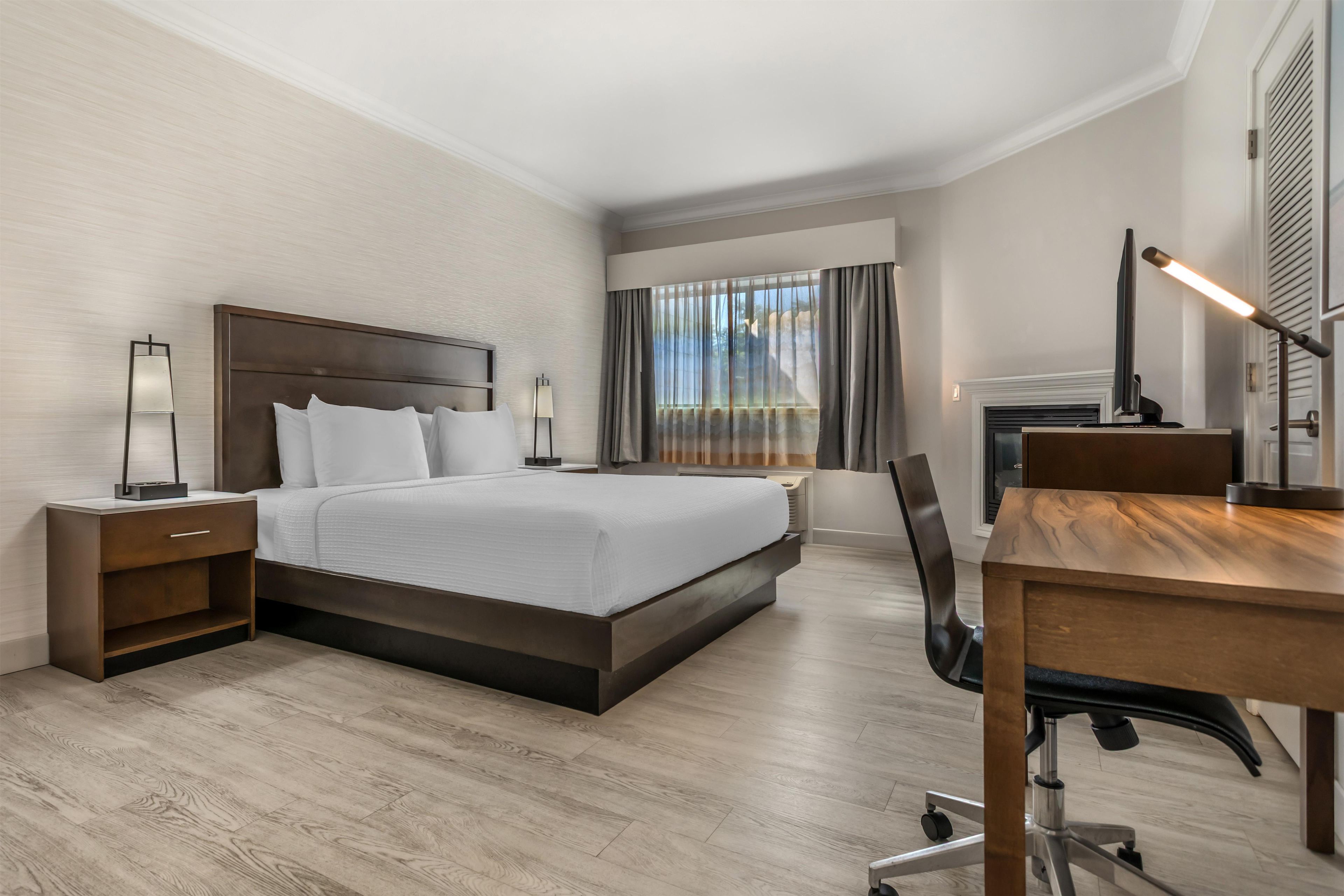 Habitació d'hotel amb un llit gran, una taula de treball, una cadira, una tauleta de nit, una làmpada i una finestra amb cortines.