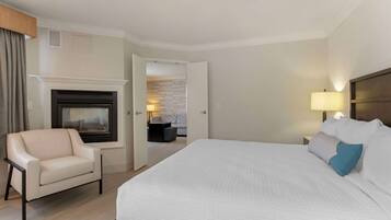 A hotel room with a large bed, a chair, a fireplace, and a nightstand with a lamp.
