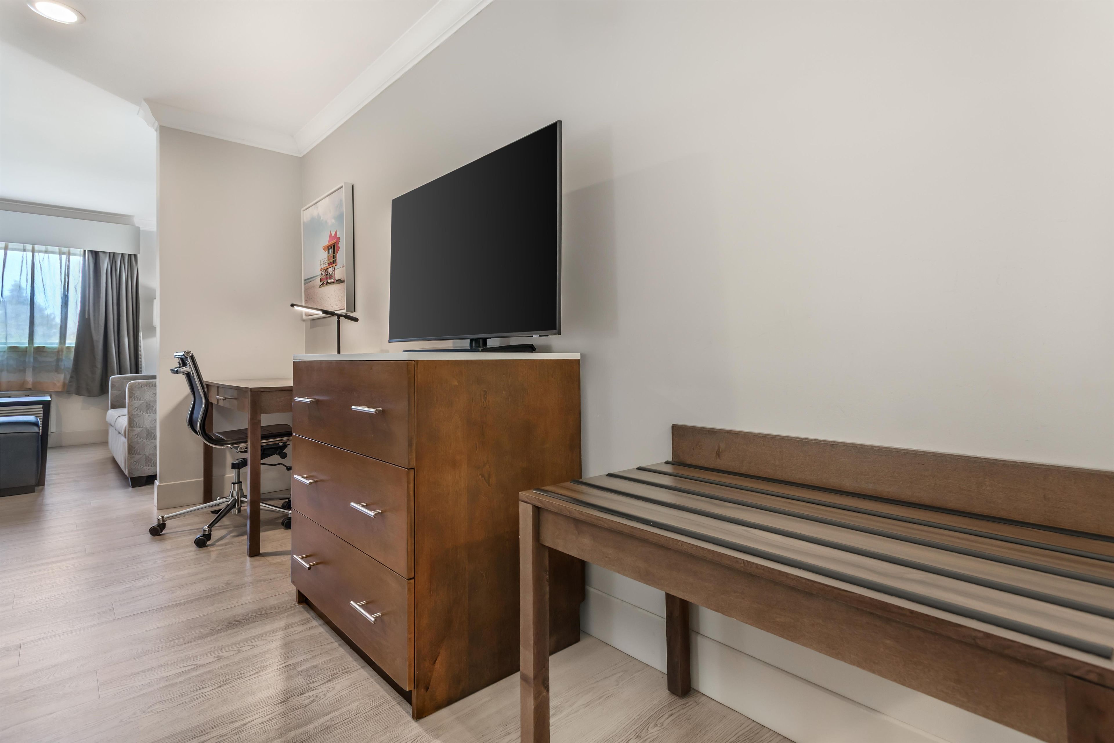 A hotel room with a wooden bench, a TV on a stand, a desk with a chair, and a large window with curtains.
