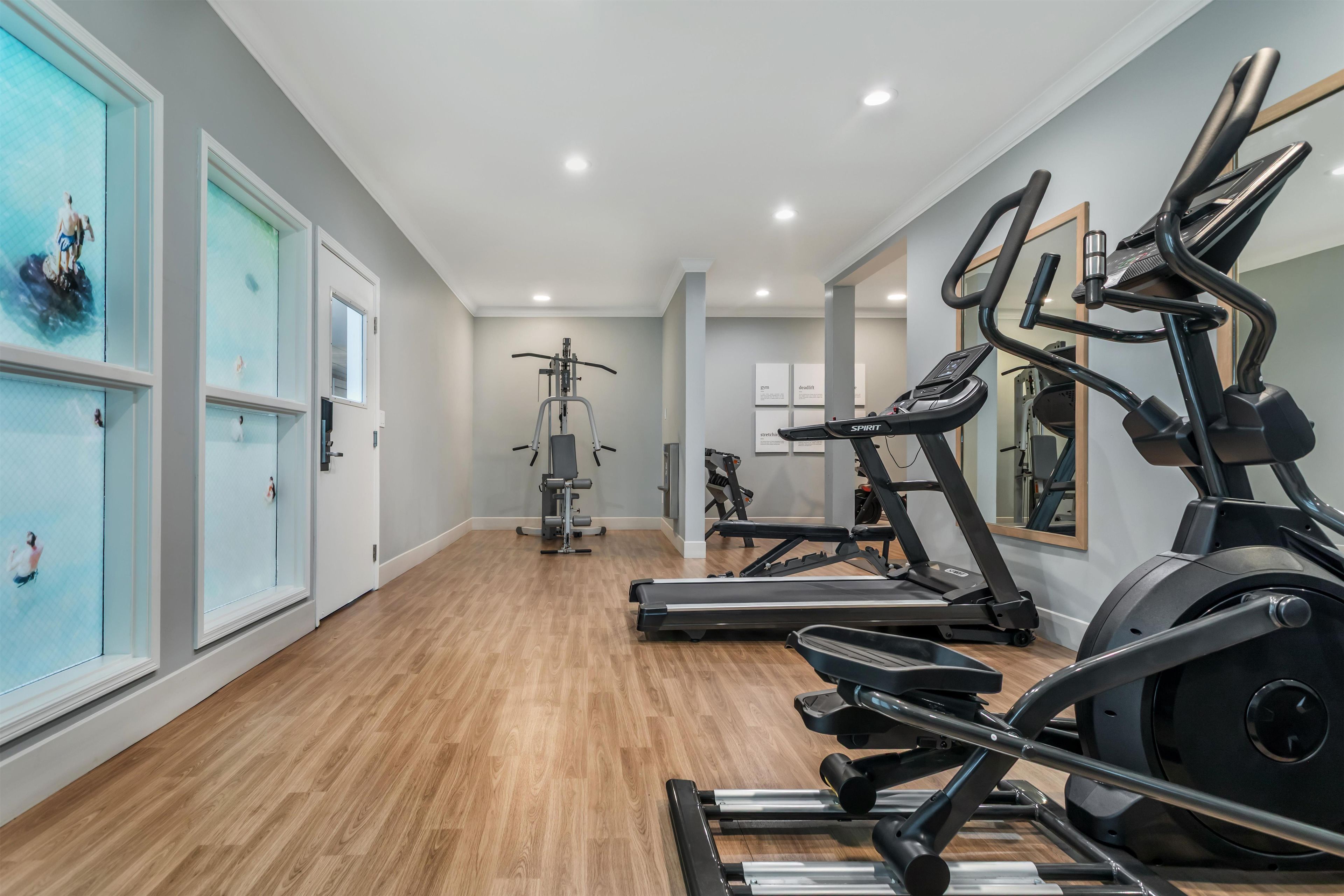 Un gimnasio con diverso equipo de ejercicio, incluyendo cintas de correr y una máquina de pesas, con piso de madera y ventanas amplias.