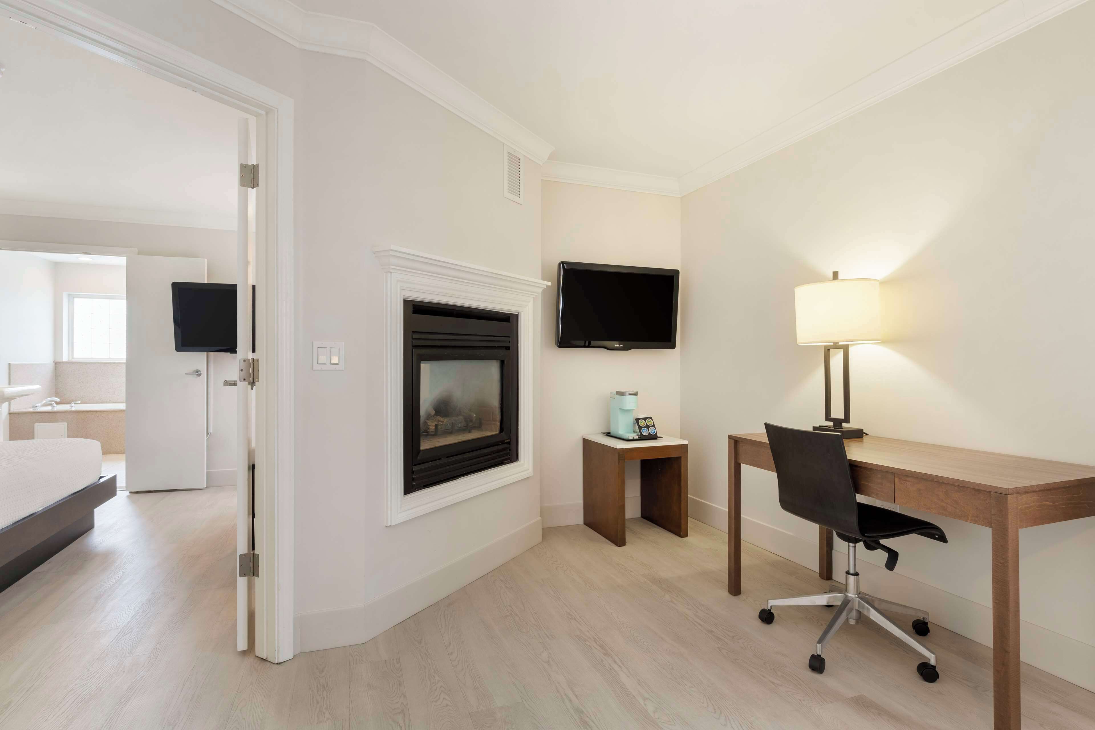A hotel room with a bed, a desk with a chair, a television, a lamp, and a fireplace.