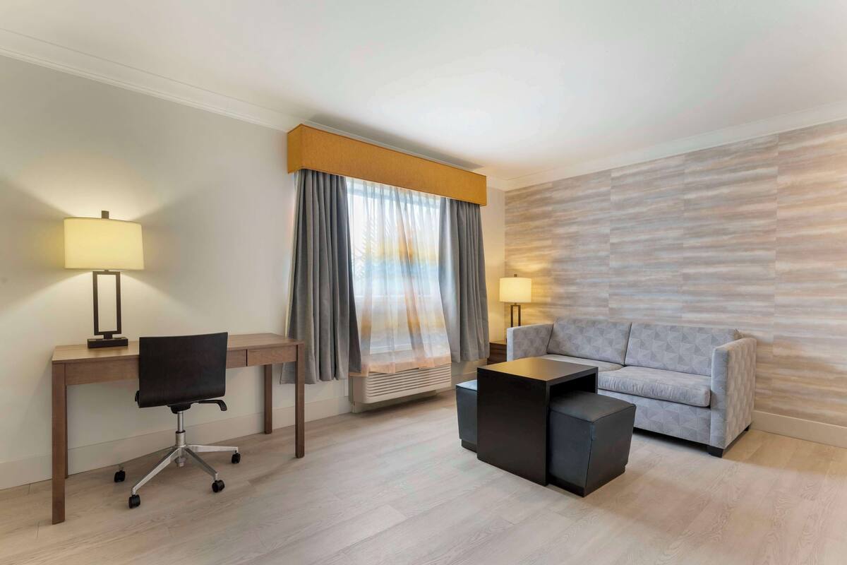 Ein modernes Hotelzimmer mit einem Holztisch, einem schwarzen Bürostuhl, einem grauen Sofa und einem kleinen schwarzen Couchtisch.