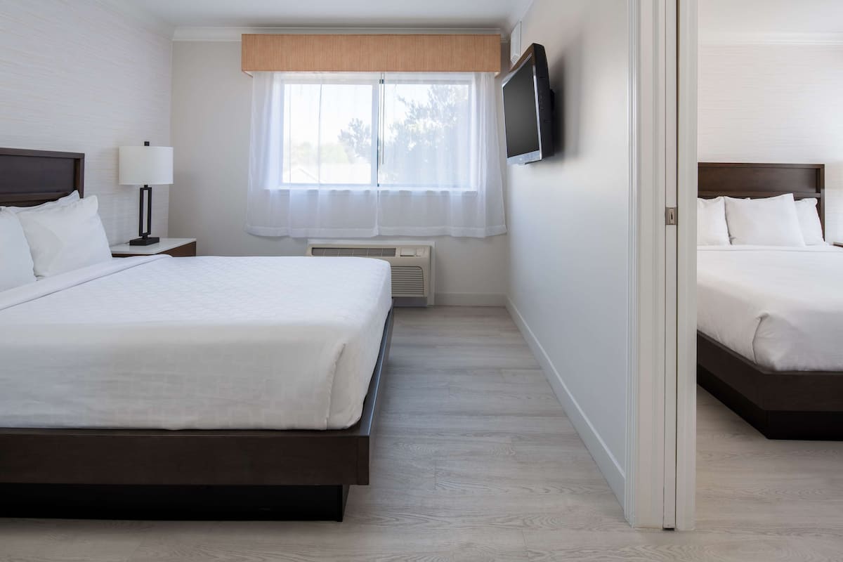 Ein Hotelzimmer mit zwei Betten, einem an der Wand befestigten Fernseher, einem Fenster mit Vorhängen und einem Nachttisch mit Lampe.