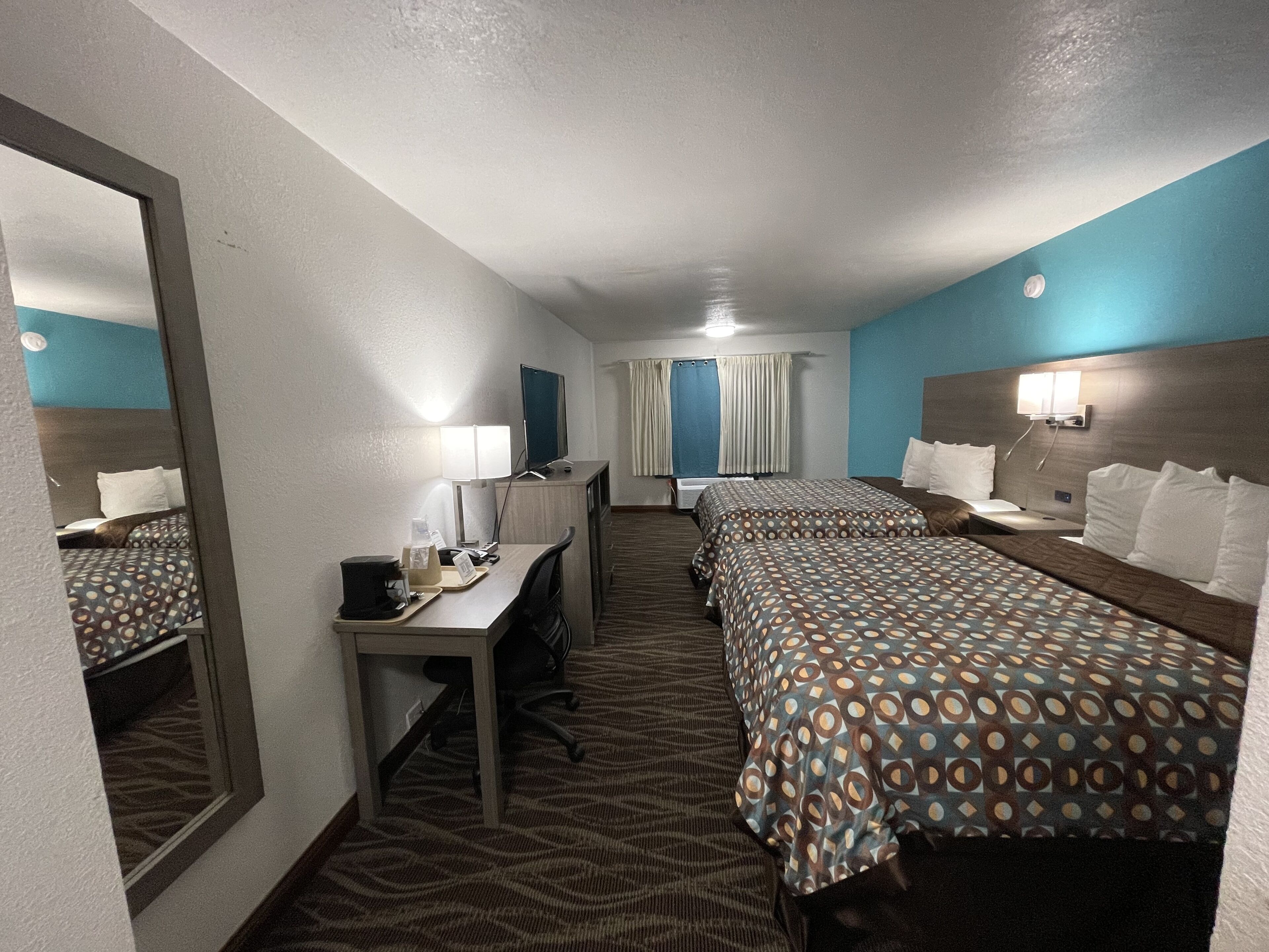 Quarto de hotel com duas camas, uma escrivaninha, uma cadeira, um espelho e uma janela com cortinas.