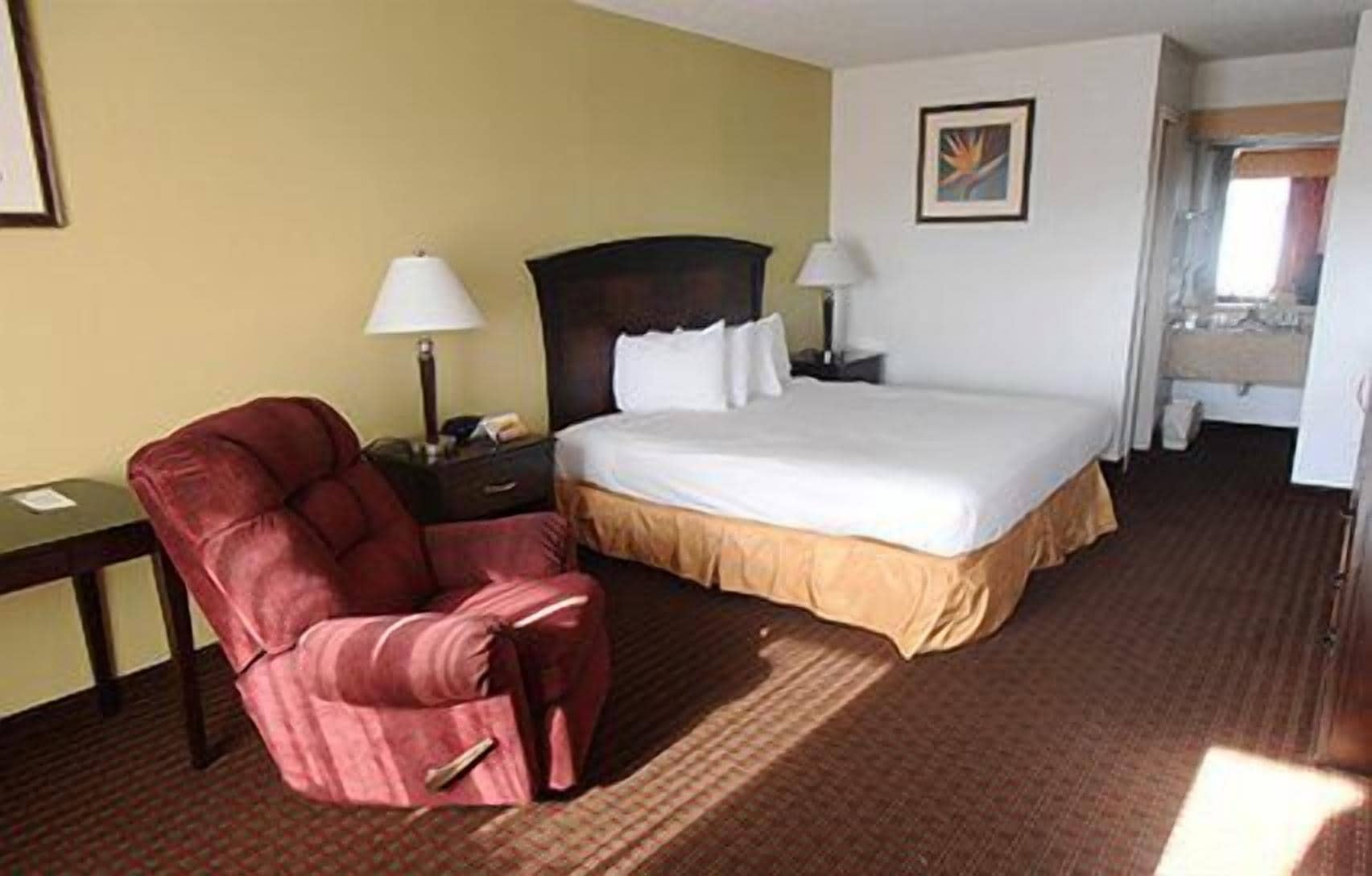 A hotel room with a bed, a red armchair, two bedside tables with lamps, and a painting on the wall.