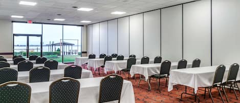 Un salón de conferencias con filas de mesas y sillas, amplias ventanas con vistas a un parque y una alfombra estampada.
