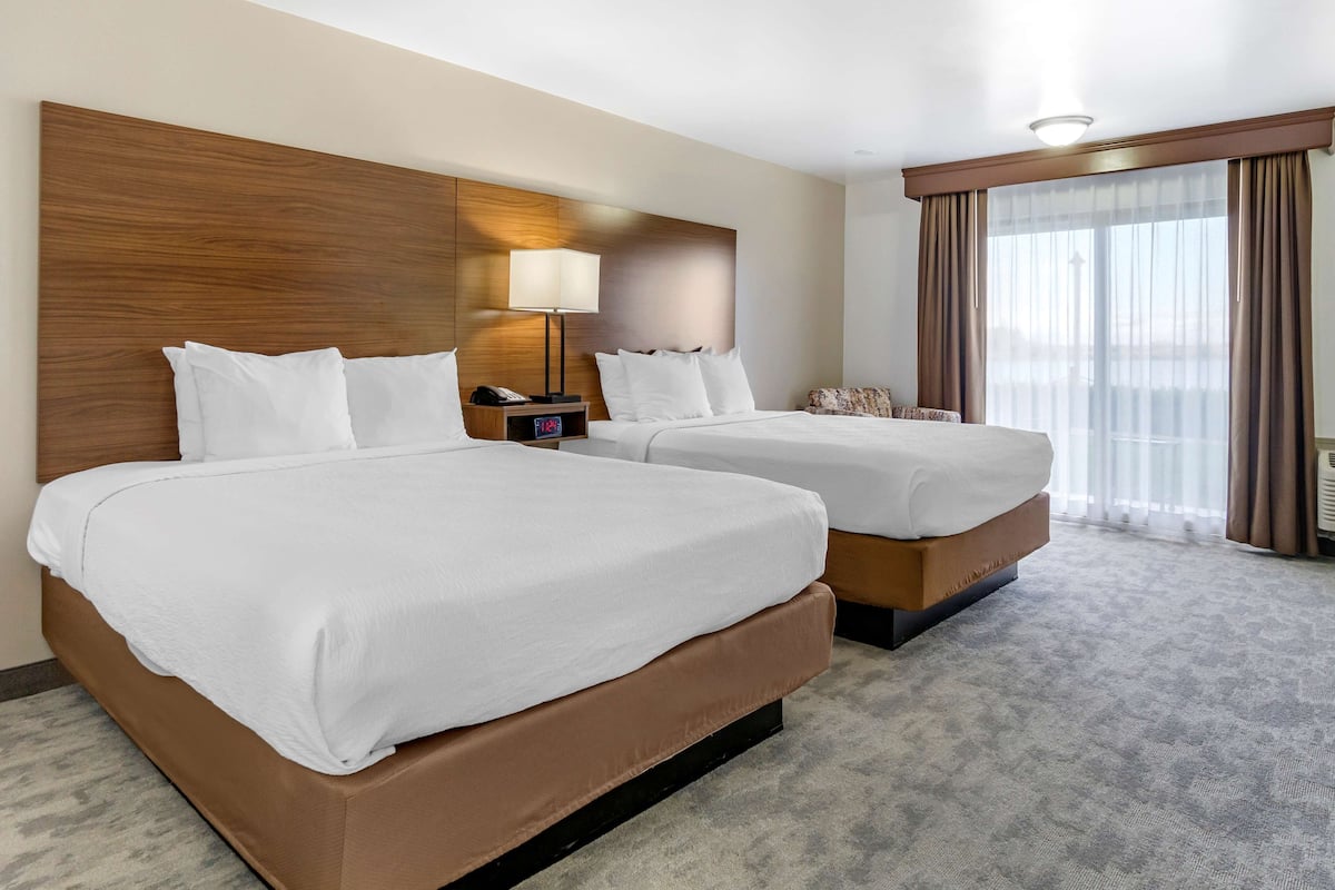 A hotel room with two beds, a nightstand with a lamp, a large window with curtains, and a view of the city.