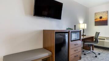 Ein Hotelzimmer mit Flachbildfernseher an der Wand, einem Holztisch mit Stuhl, einer Bank und einem Wandbild eines Sonnenuntergangs.