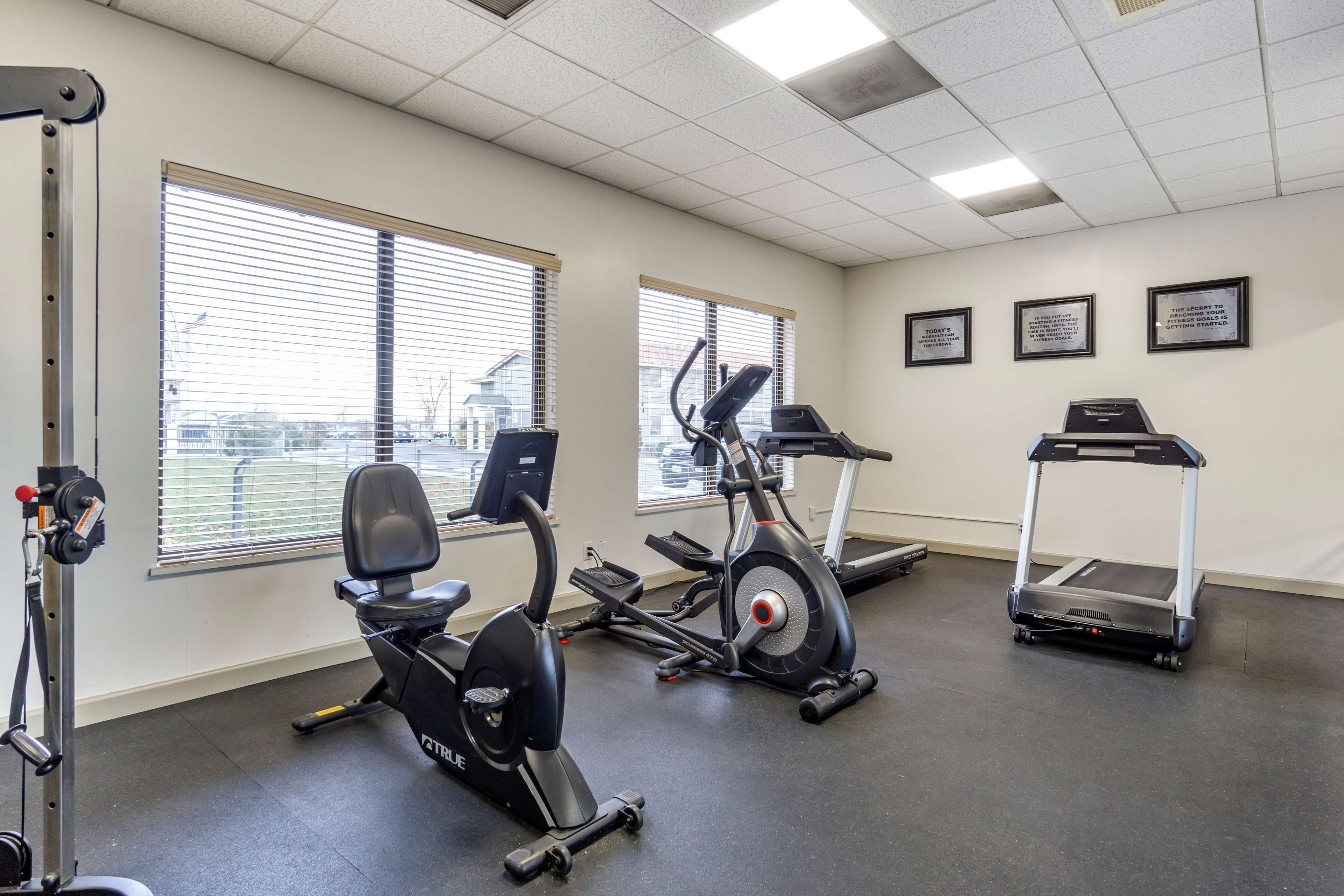 Ein Fitnessstudio mit Trainingsgeräten, darunter ein Crosstrainer, ein Laufband und ein Ergometer. Große Fenster mit Jalousien und motivierende Poster schmücken die Wände.