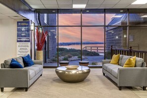 A modern lobby with two sofas, a central coffee table, and a large window offering a view of a sunset over a body of water. - Best Western Plus Lake Front Hotel (Moses Lake)