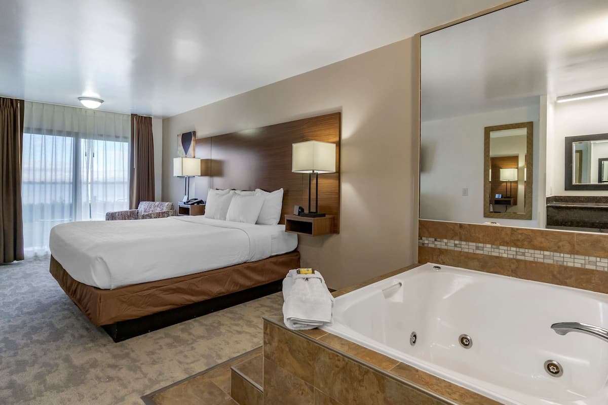 A hotel room with a large bed, a separate bathroom with a bathtub, a mirror, and a window with curtains.