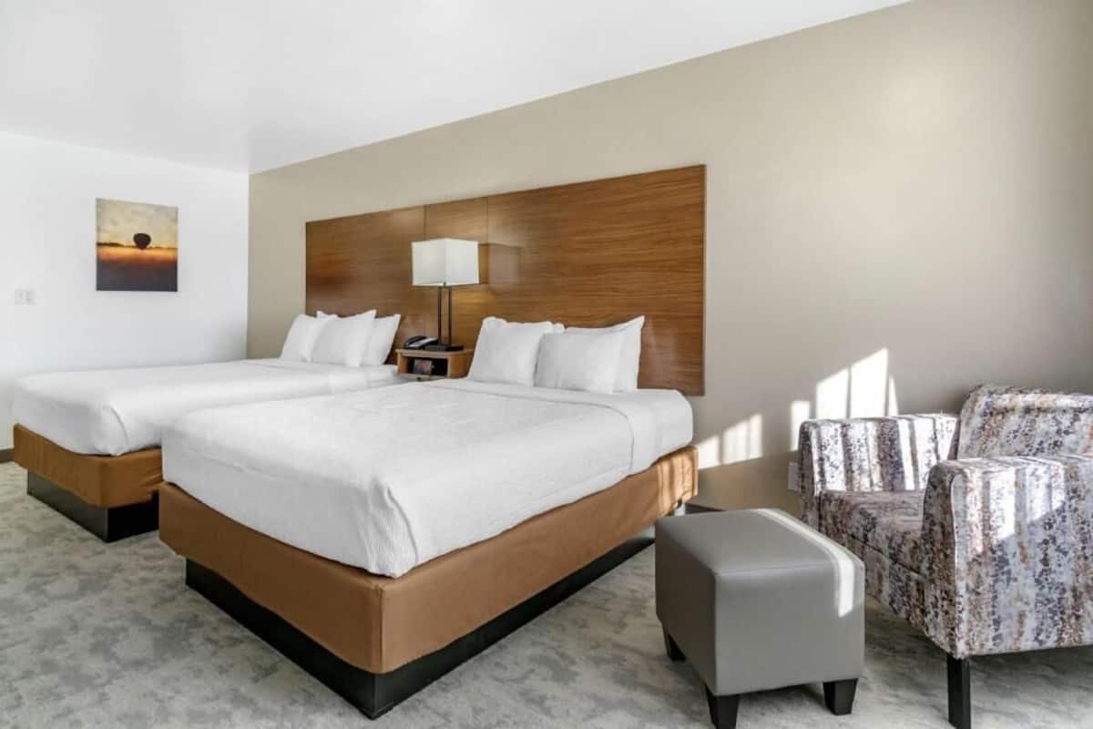 A hotel room with two beds, a nightstand, a lamp, a chair, and a painting on the wall.