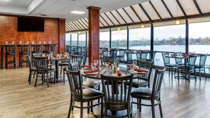Un restaurante amplio con numerosas mesas y sillas, amplias ventanas que ofrecen vistas a un lago y un televisor de pantalla plana montado en la pared.