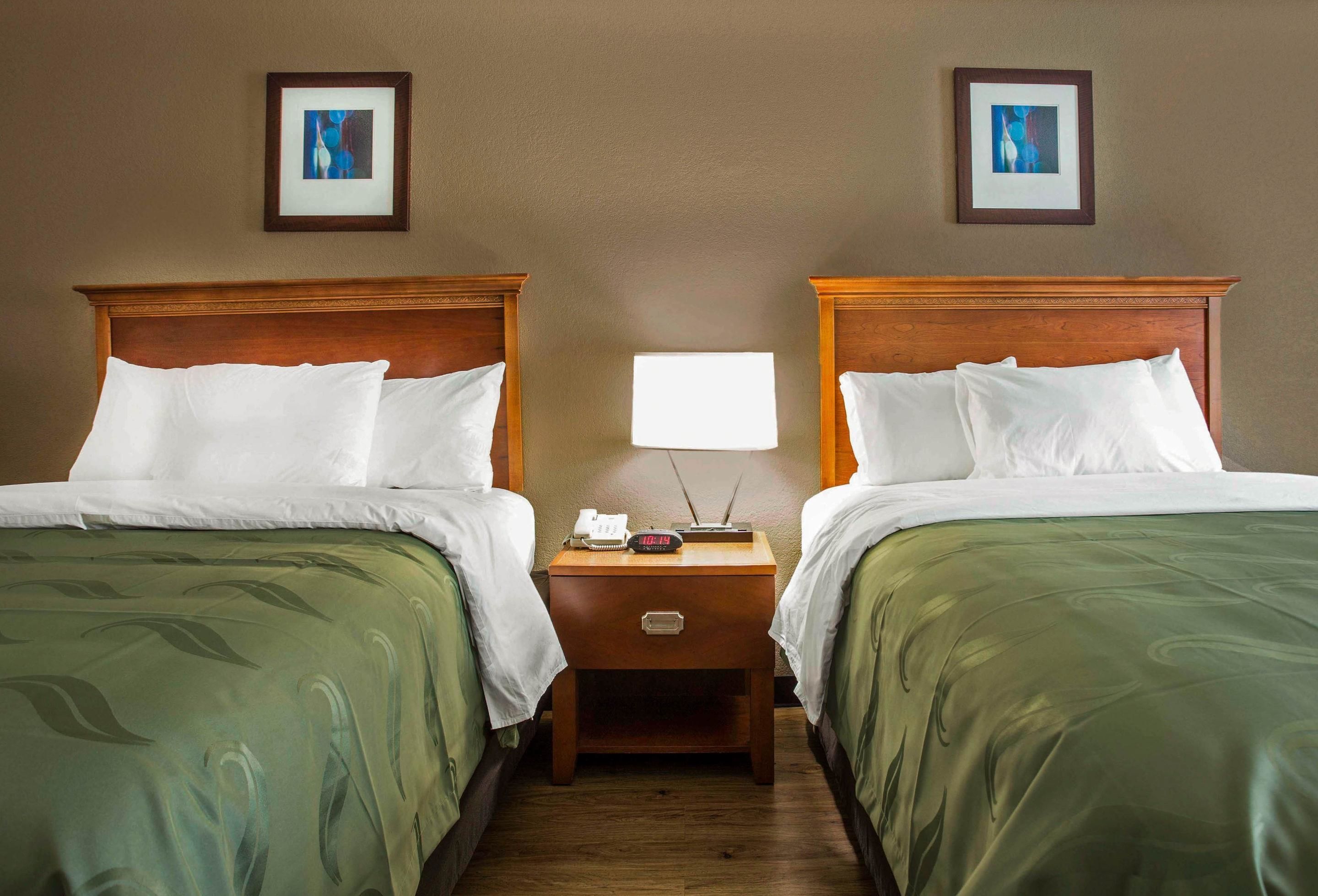 Une chambre d’hôtel avec deux lits, chacun doté d’une tête de lit en bois, d’une table de chevet avec une lampe et d’un petit tiroir.
