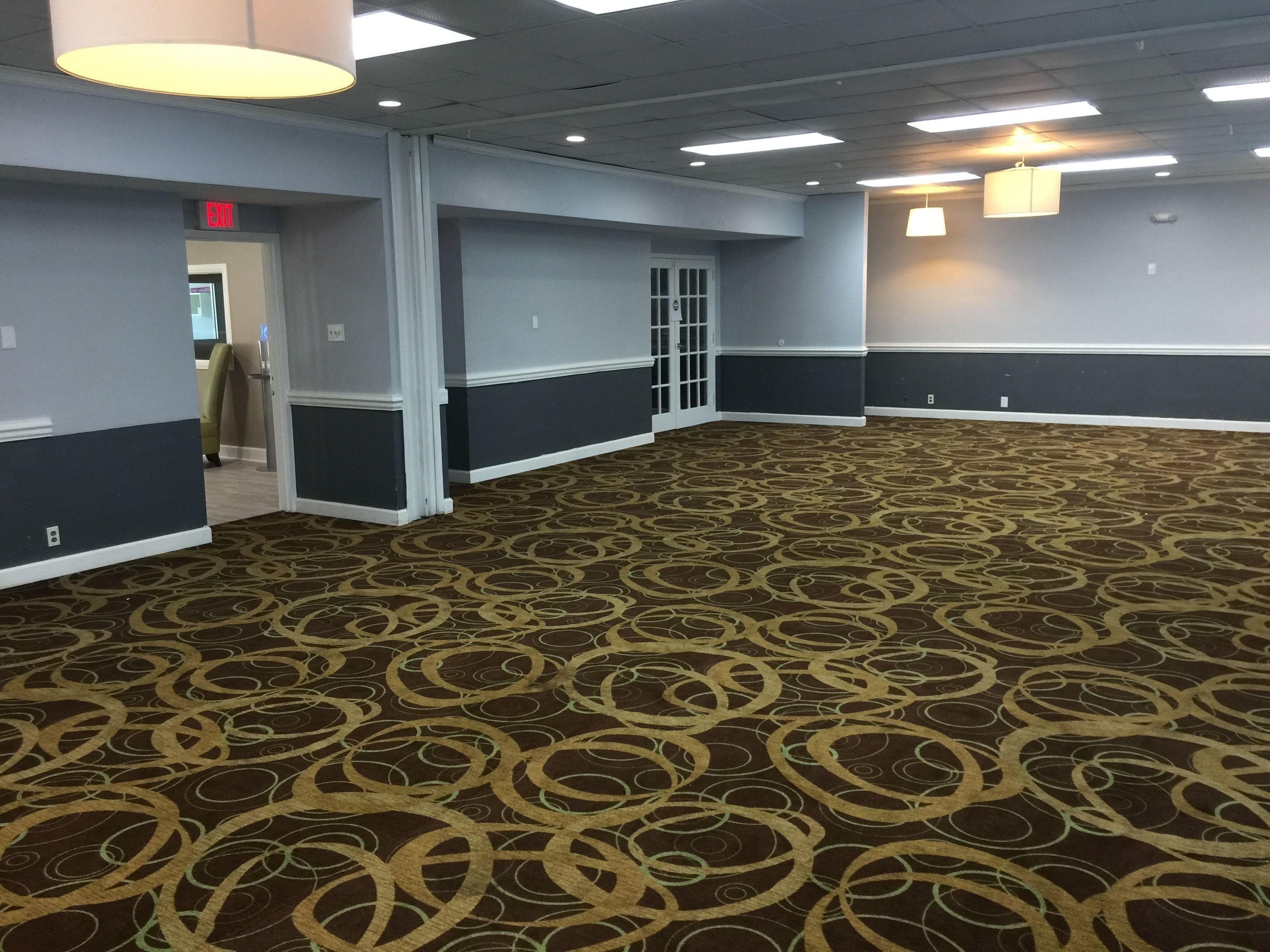 A spacious hallway with patterned carpet, a door with a glass panel, and a red exit sign.