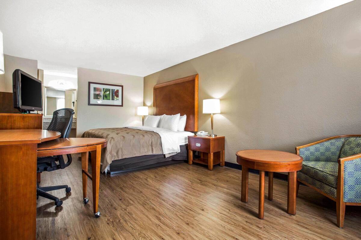 A hotel room with a bed, bedside table, lamp, chair, desk, and a television.