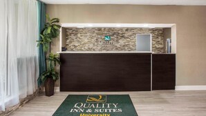 Ein Empfangsbereich mit dunklem Empfangstresen, einem Logo und einer grünen Fußmatte mit der Aufschrift 'Quality Inn & Suites University'.