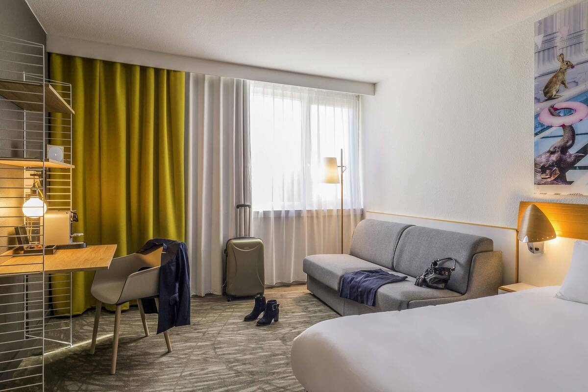 Une chambre d’hôtel avec un lit, un canapé, un bureau avec une lampe, une chaise, une valise et un tableau au mur.