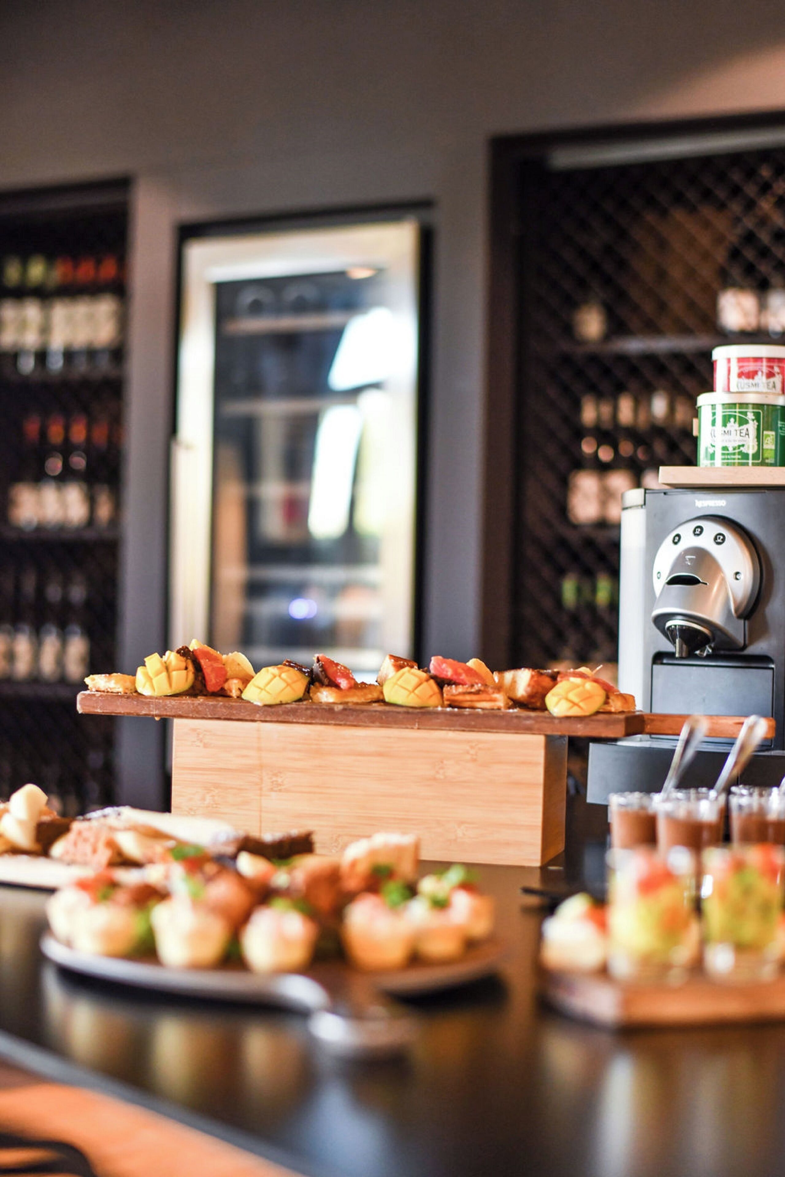 A bar area with a variety of food items on a wooden board, a coffee machine, and a wine cooler in the background.