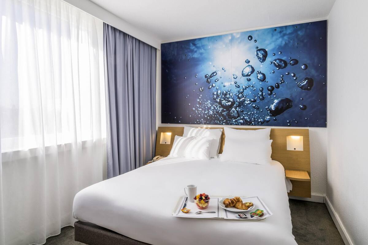 Une chambre d’hôtel avec un grand lit, un plateau de petit-déjeuner avec des croissants et des fruits, un tableau représentant des gouttes d’eau sur le mur et une fenêtre avec des rideaux.