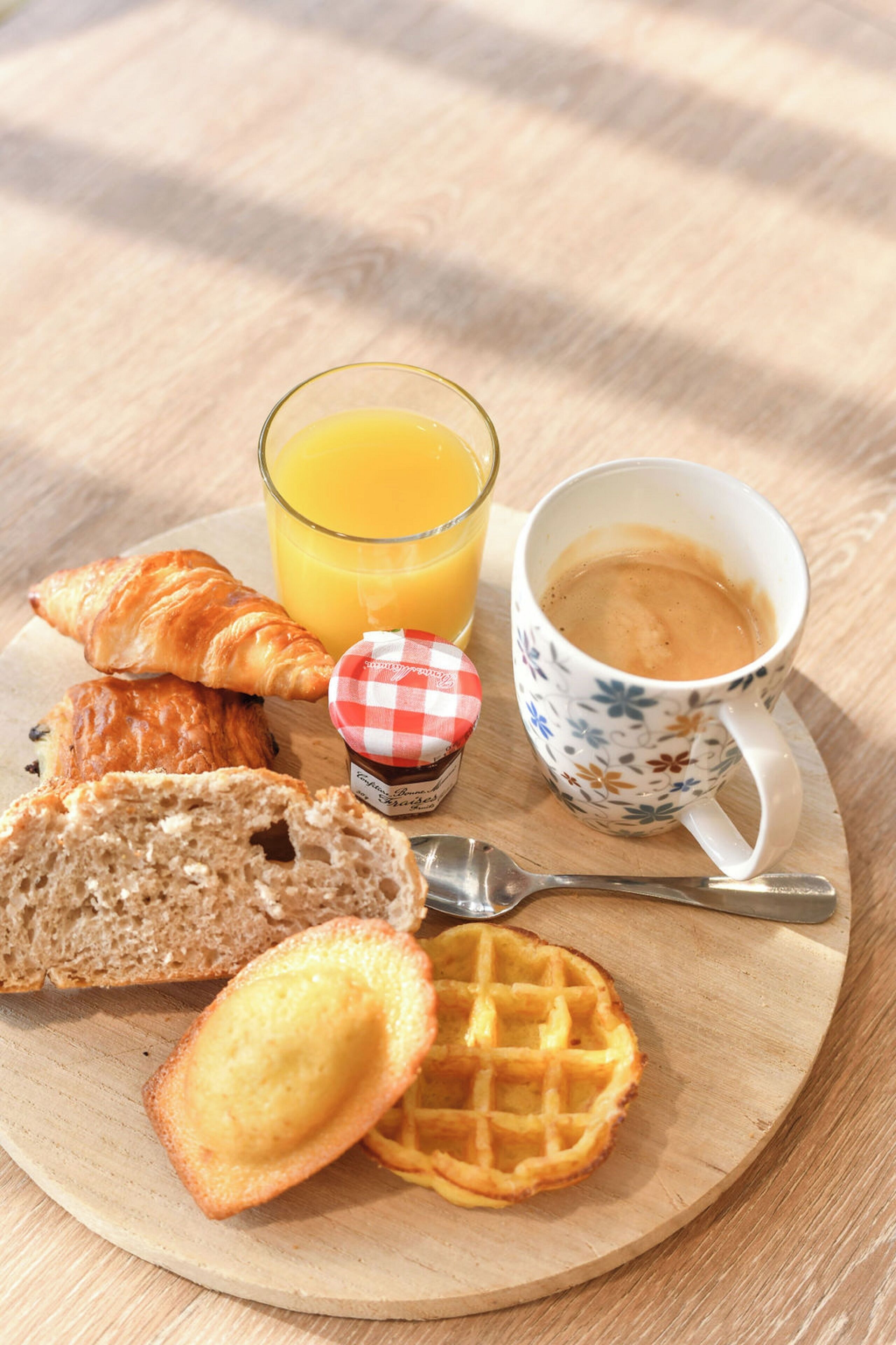 Une assiette de petit-déjeuner avec des croissants, du pain, une gaufre, un verre de jus d’orange et une tasse de café, le tout posé sur une table en bois.