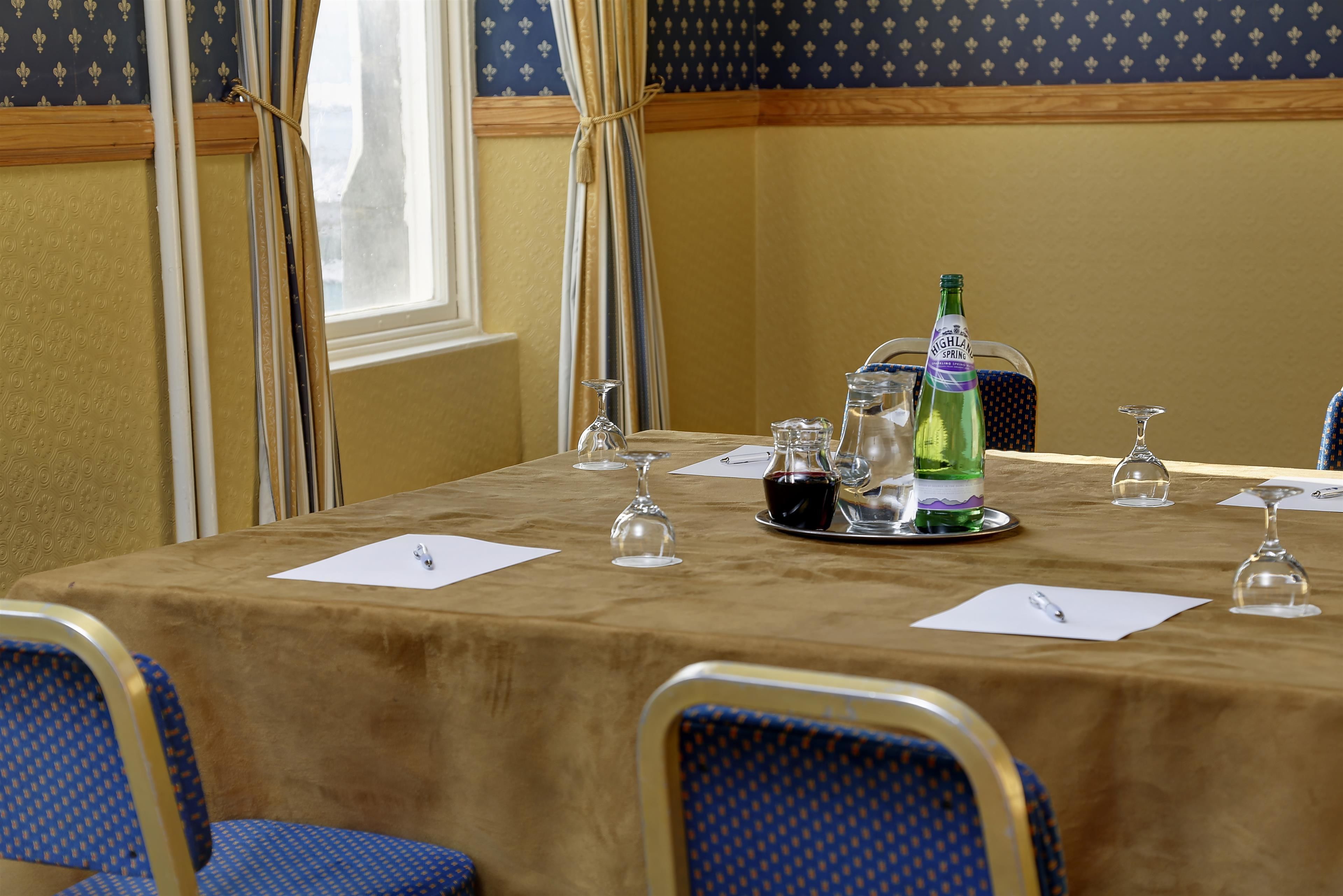 Une salle de conférence avec une table préparée pour une réunion, comprenant une bouteille d’eau, des verres à vin et un bloc-notes.