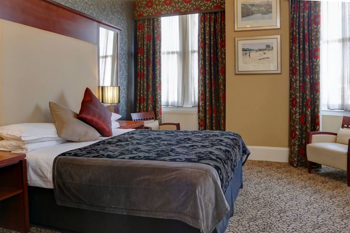 A hotel room with a large bed, a bedside table, a chair, and a window with curtains.