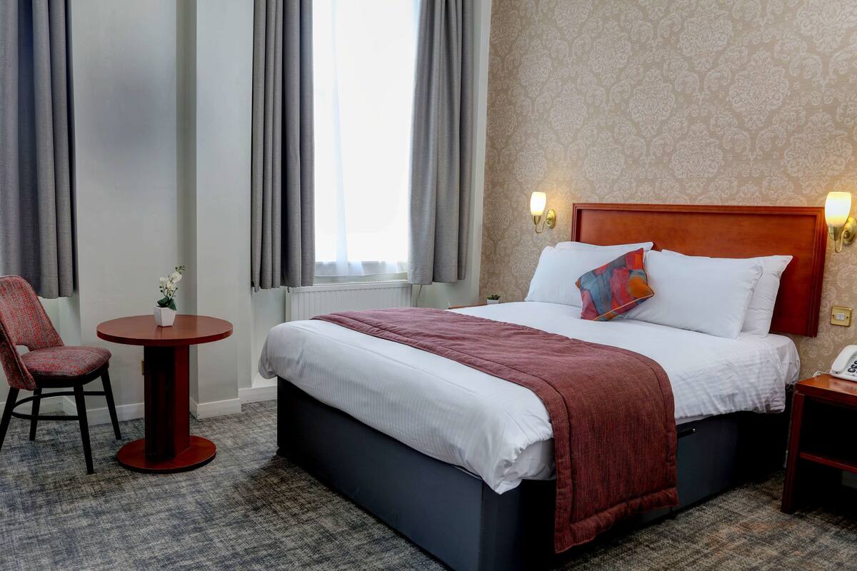 A hotel room with a bed, a small round table, a chair, and a nightstand with a phone.