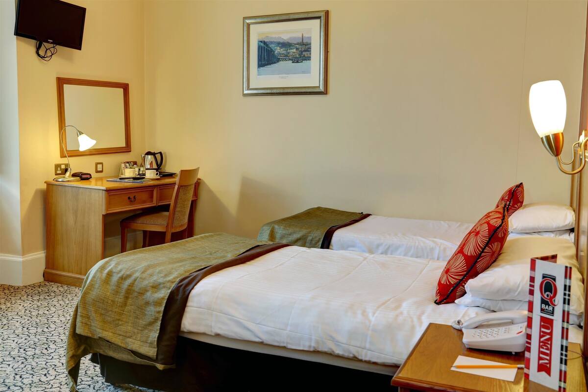 A hotel room with two beds, a desk with a chair, a desk lamp, a framed picture on the wall, and a bedside table with a phone and a book.