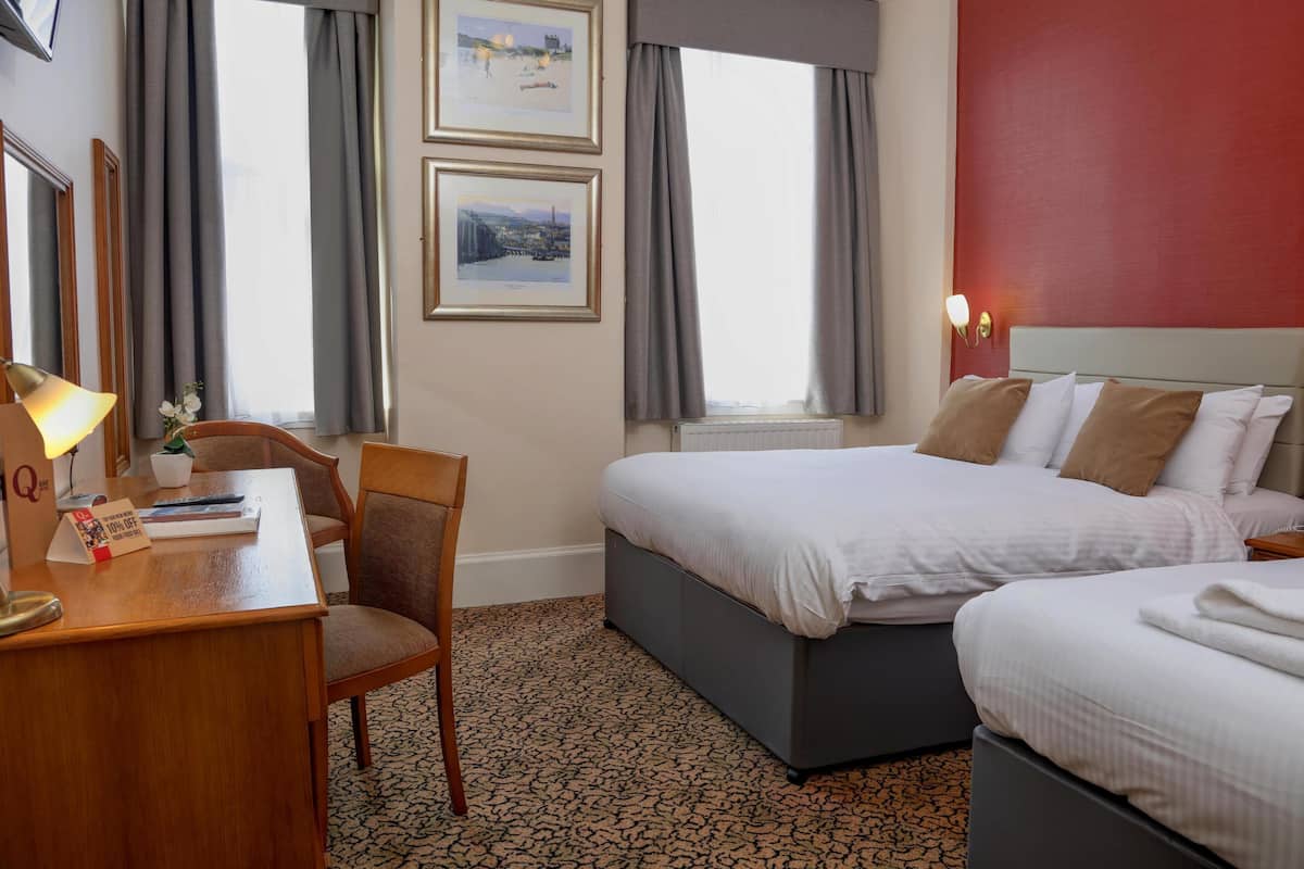 A hotel room with a desk, chair, two beds, a window with curtains, and framed pictures on the wall.
