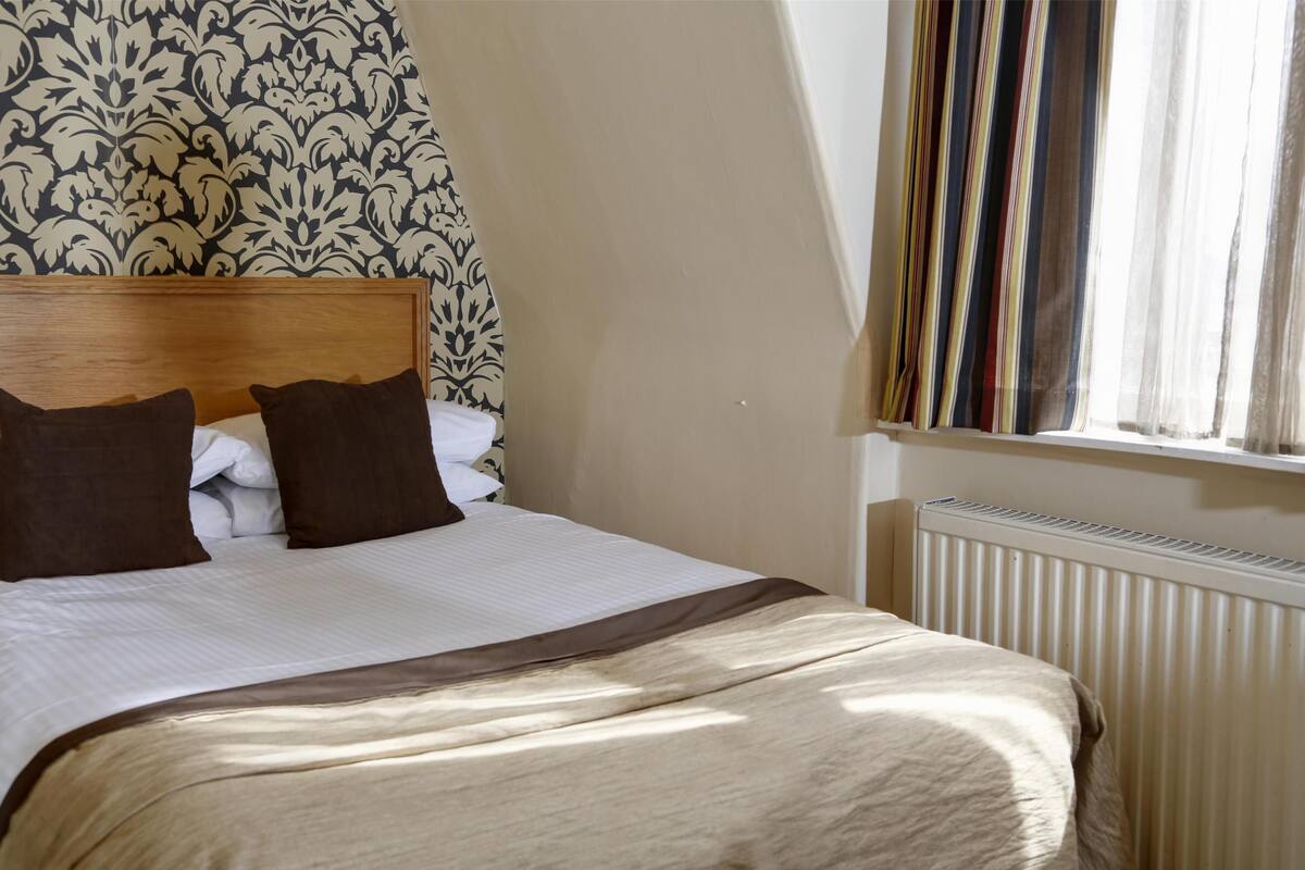A neatly made bed with a wooden headboard, a striped window blind, and a radiator.