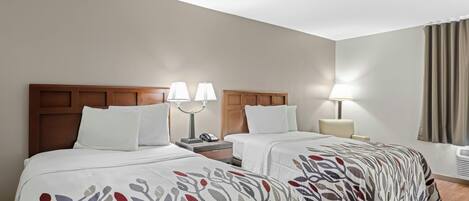 A hotel room with two beds, each with a wooden headboard, white bedding, and a patterned throw. There are bedside tables with lamps and a phone.